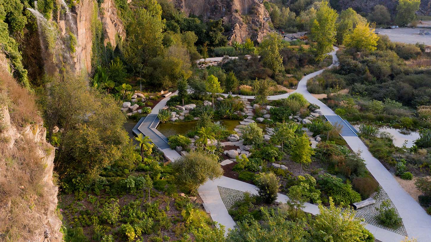 Vue aérienne d'un parc urbain paysager avec sentiers sinueux, bassins et végétation luxuriante au pied d'une falaise