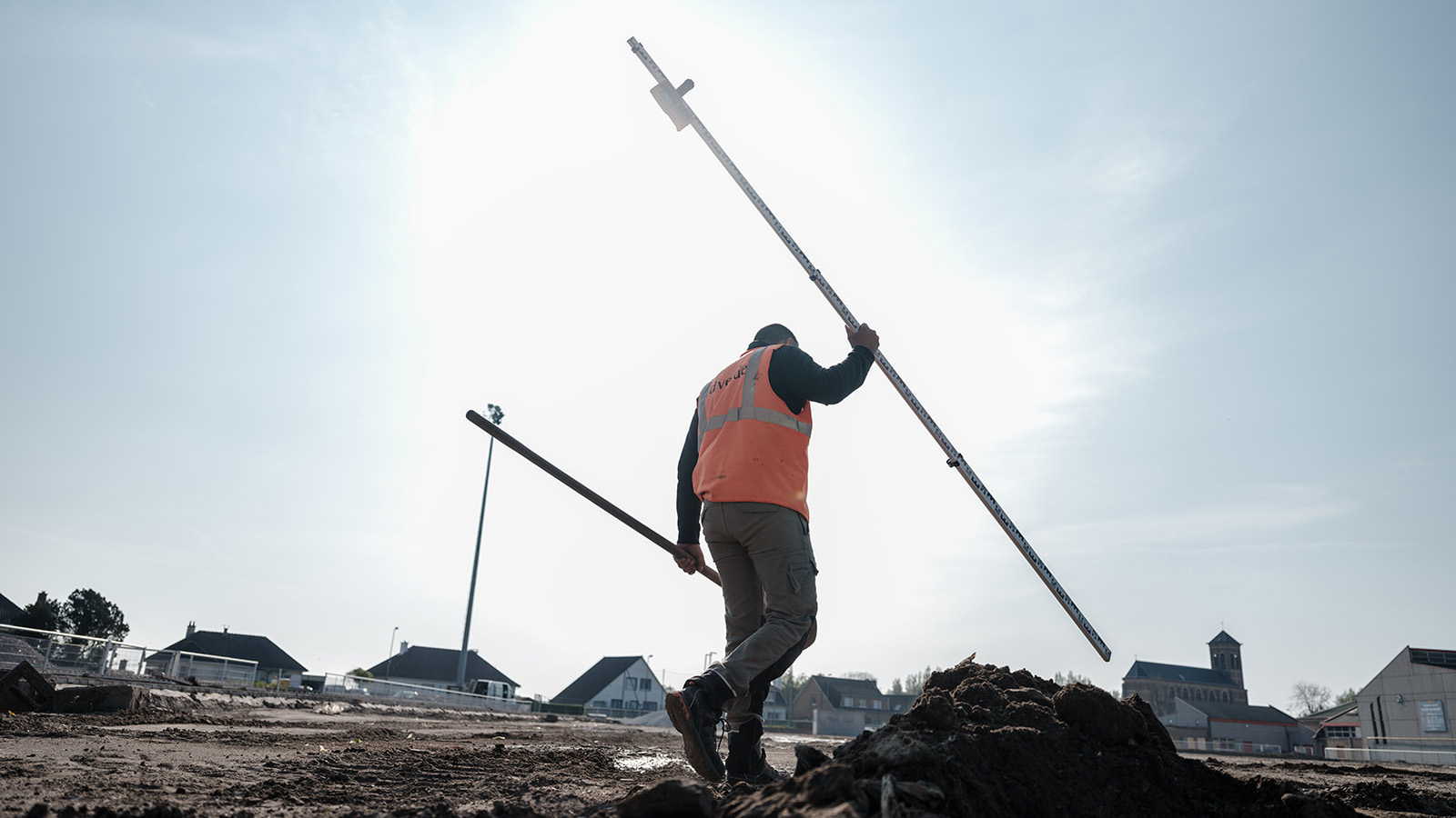 Ouvrier idverde en gilet orange portant une mire de nivellement sur un chantier de terrassement