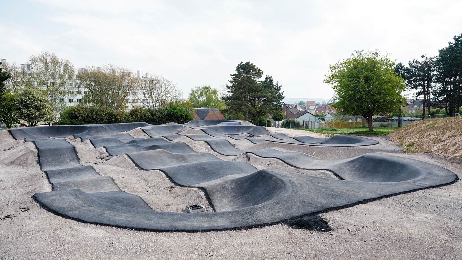 Piste de pump track en asphalte noir avec bosses et virages, entourée d'arbres et de bâtiments urbains