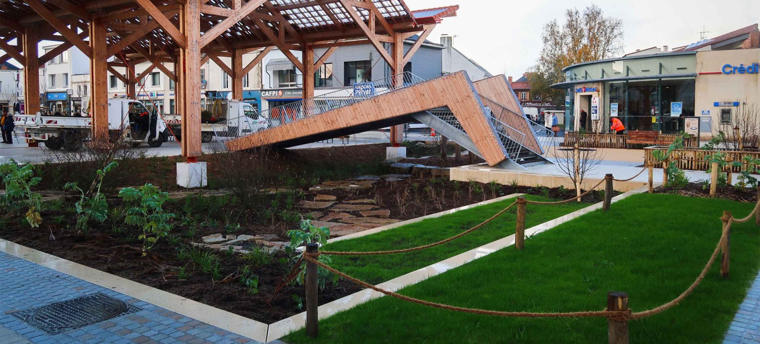 Aménagement paysager avec pelouse, massifs plantés et passerelle en bois sur une place de centre-ville, à Challans