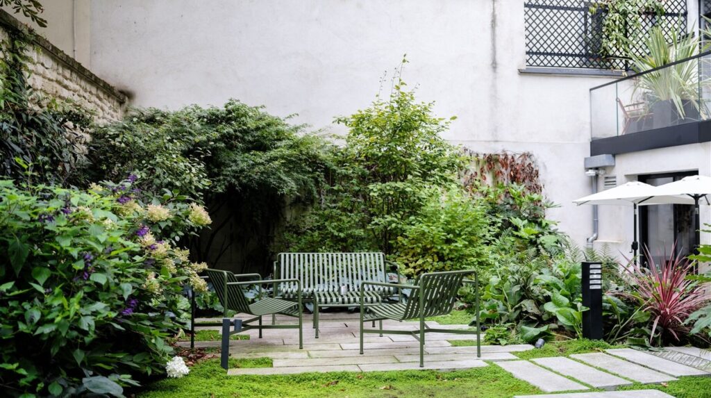 Jardin de ville clos avec terrasse, bancs et végétation dense contre un mur blanc