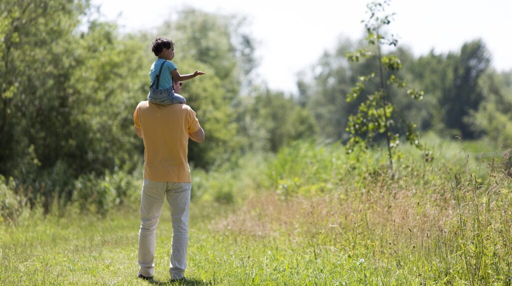 Père portant son enfant sur les épaules dans une prairie haute au bord de l'eau