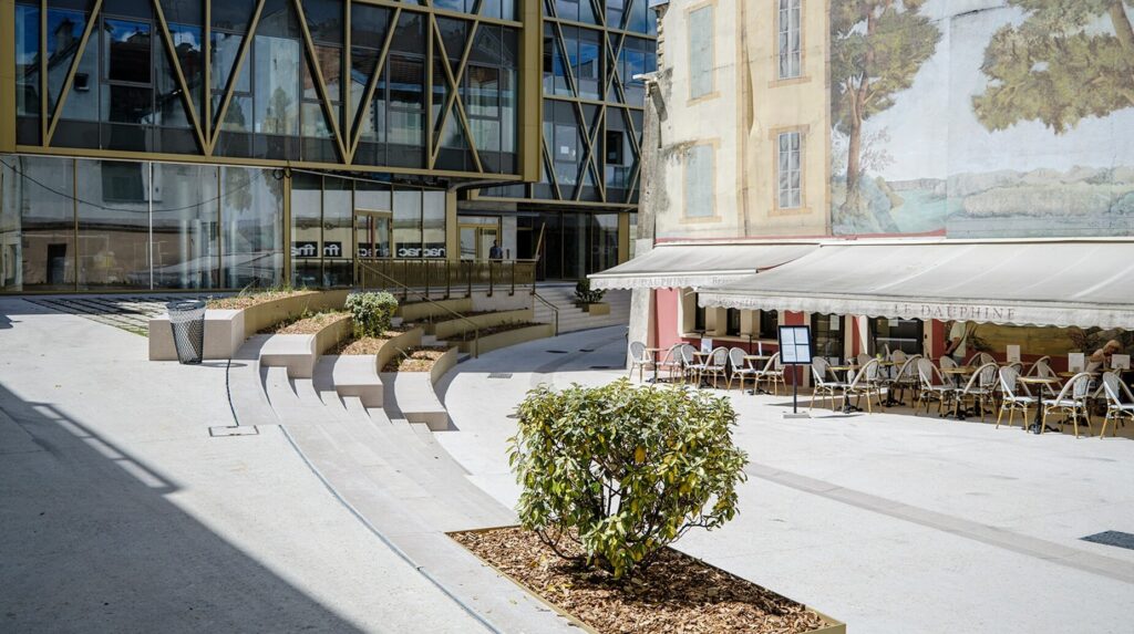 Parvis minéral avec bacs plantés devant un bâtiment contemporain vitré