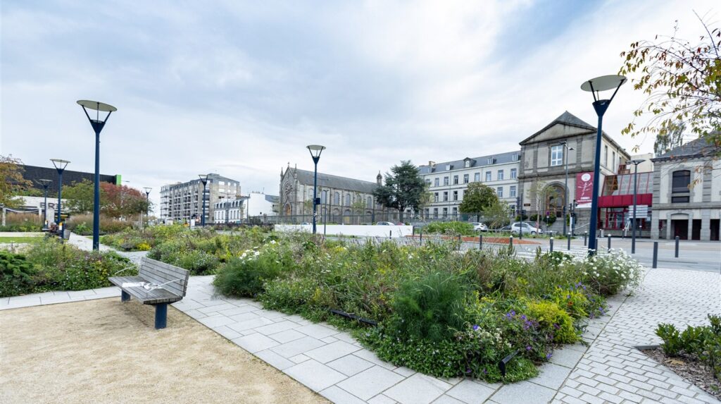 Place publique paysagée avec massifs de graminées, bancs et lampadaires devant des façades